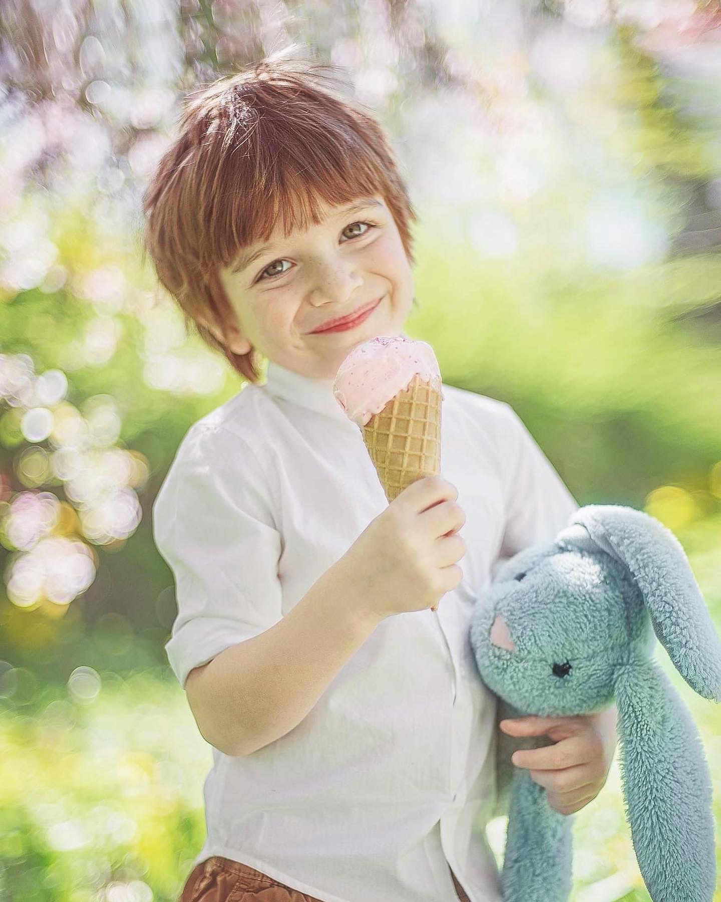 Uśmiechnięty chłopiec z brązowymi włosami trzyma w jednej ręce różowe lody w wafelku, a w drugiej pluszowego, niebieskiego królika na tle rozmytej zieleni i kwitnących drzew.