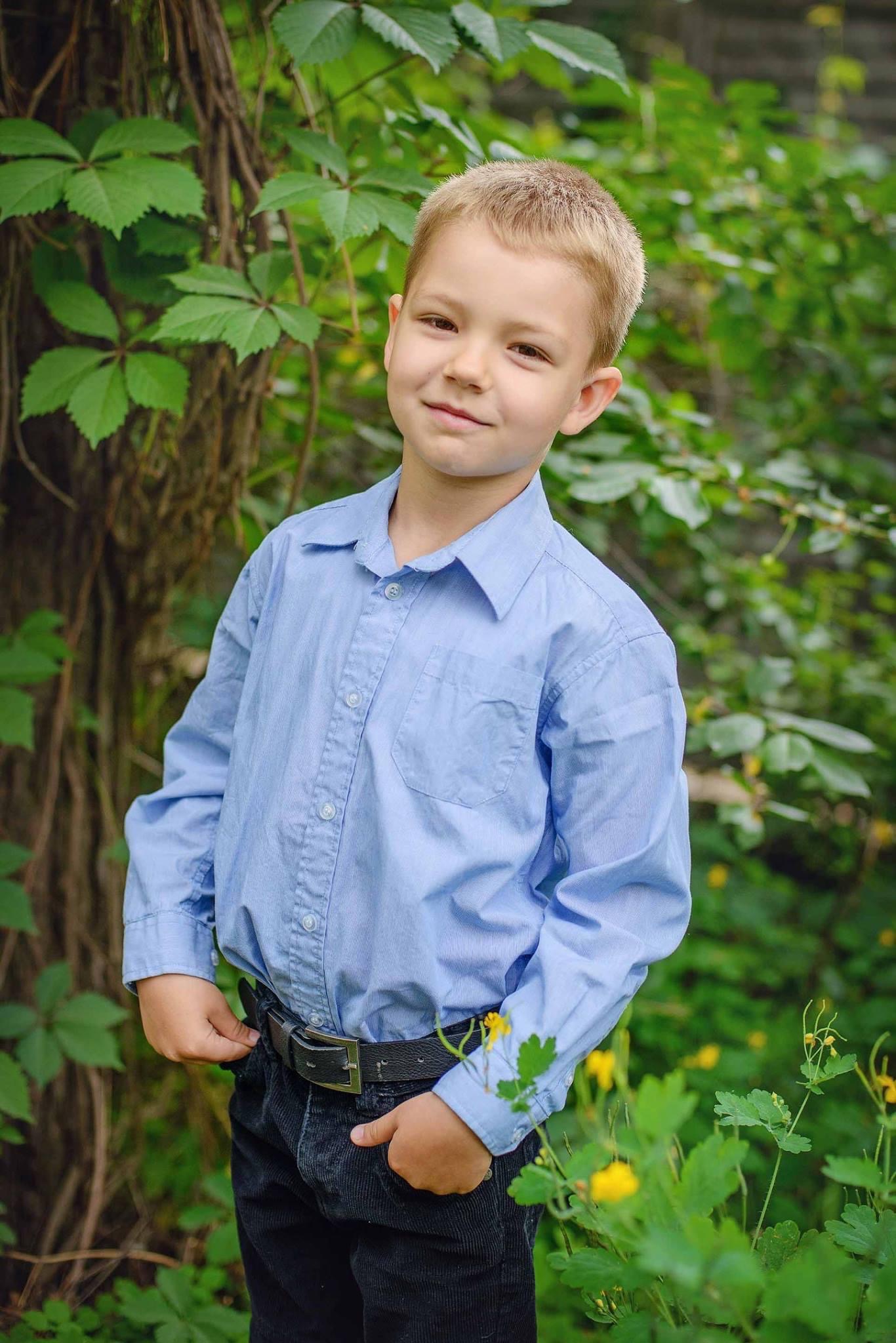 Uśmiechnięty chłopiec w błękitnej koszuli i czarnych spodniach, pozujący na tle zieleni z żółtymi kwiatami.