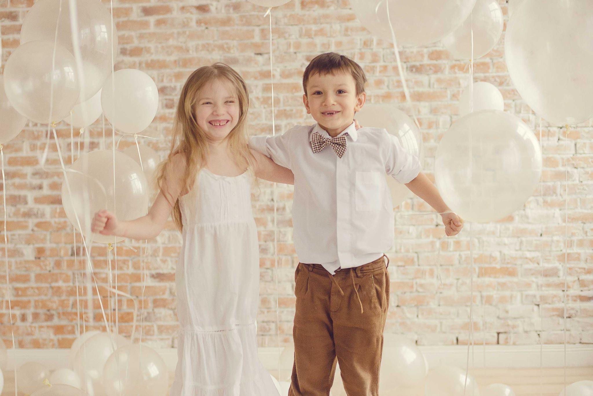 Uśmiechnięta dziewczynka w białej sukience i chłopiec z brązową muszką, obejmujący się na tle ceglanej ściany i białych balonów.