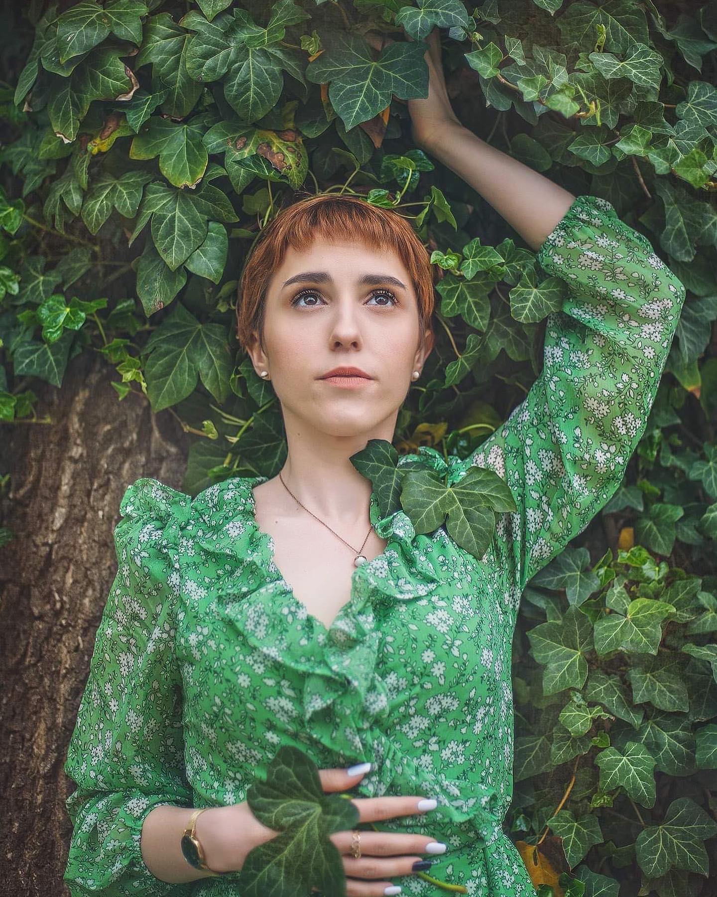 Portret kobiety z rudymi włosami w zielonej sukience w kwiaty, pozującej na tle ściany z bluszczu, spoglądającej w górę.