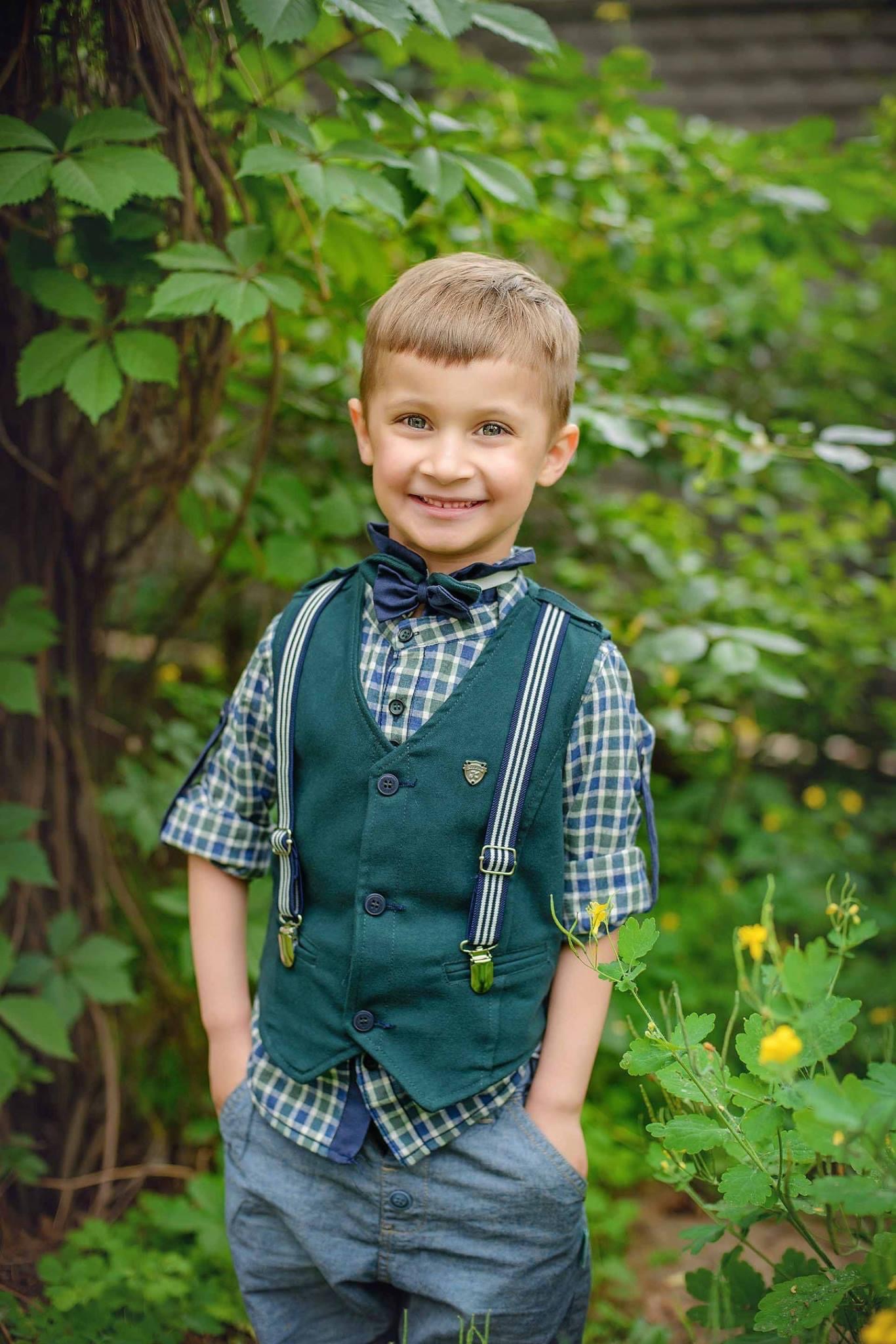 Uśmiechnięty chłopiec w zielonej kamizelce, koszuli w kratę i muszce, pozujący na tle zieleni.