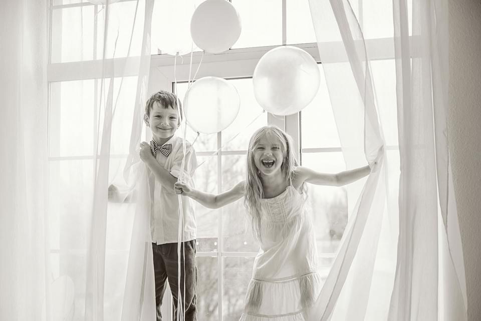 Czarno-białe zdjęcie z Lublina przedstawia uśmiechnięte rodzeństwo: chłopiec w muszce trzyma balony, a dziewczynka rozsuwa firankę, stojąc na tle okna.