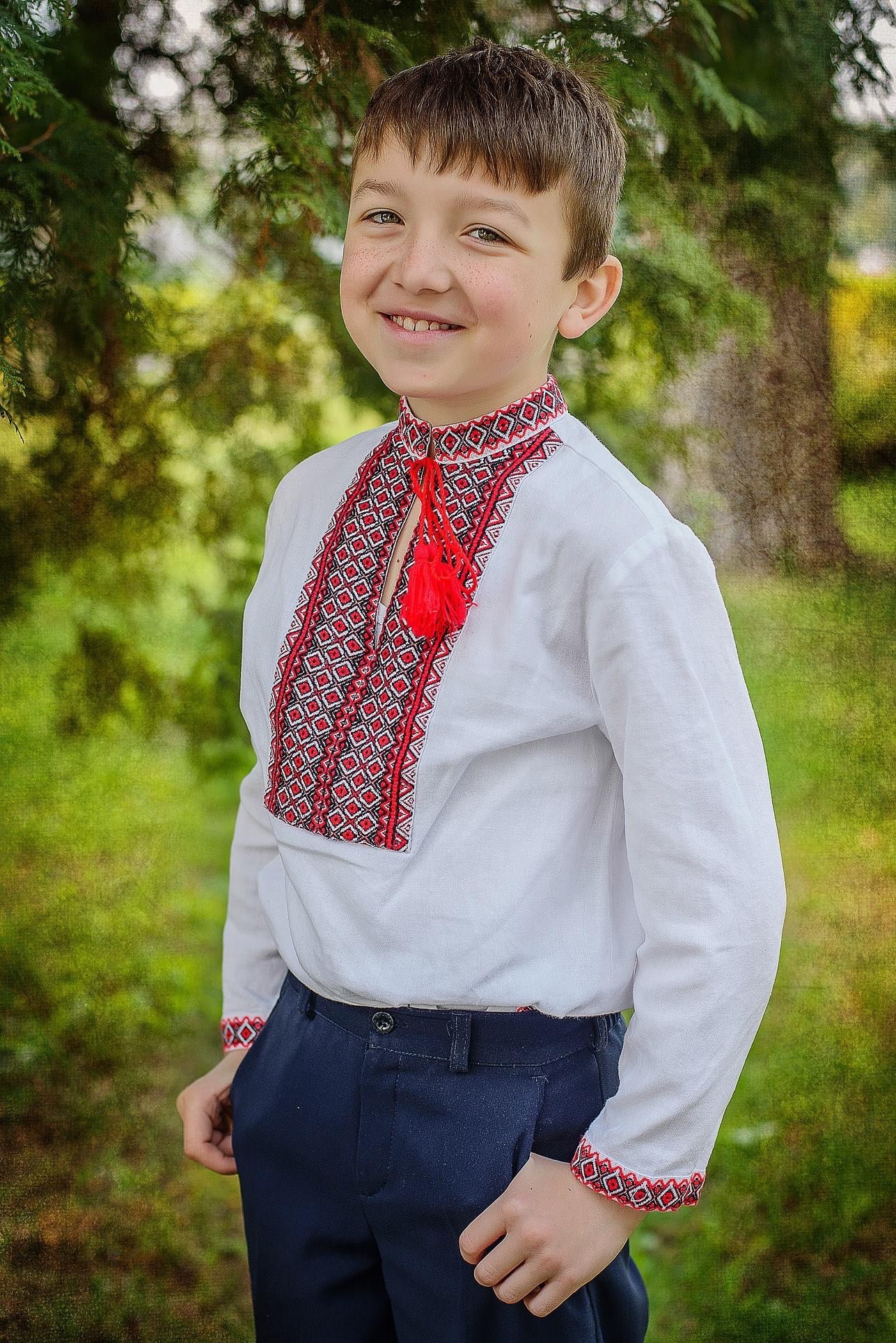 Uśmiechnięty chłopiec w białej koszuli z czerwono-czarnym haftem i ozdobnym czerwonym sznurkiem z frędzlami, pozujący na zewnątrz na tle zieleni.