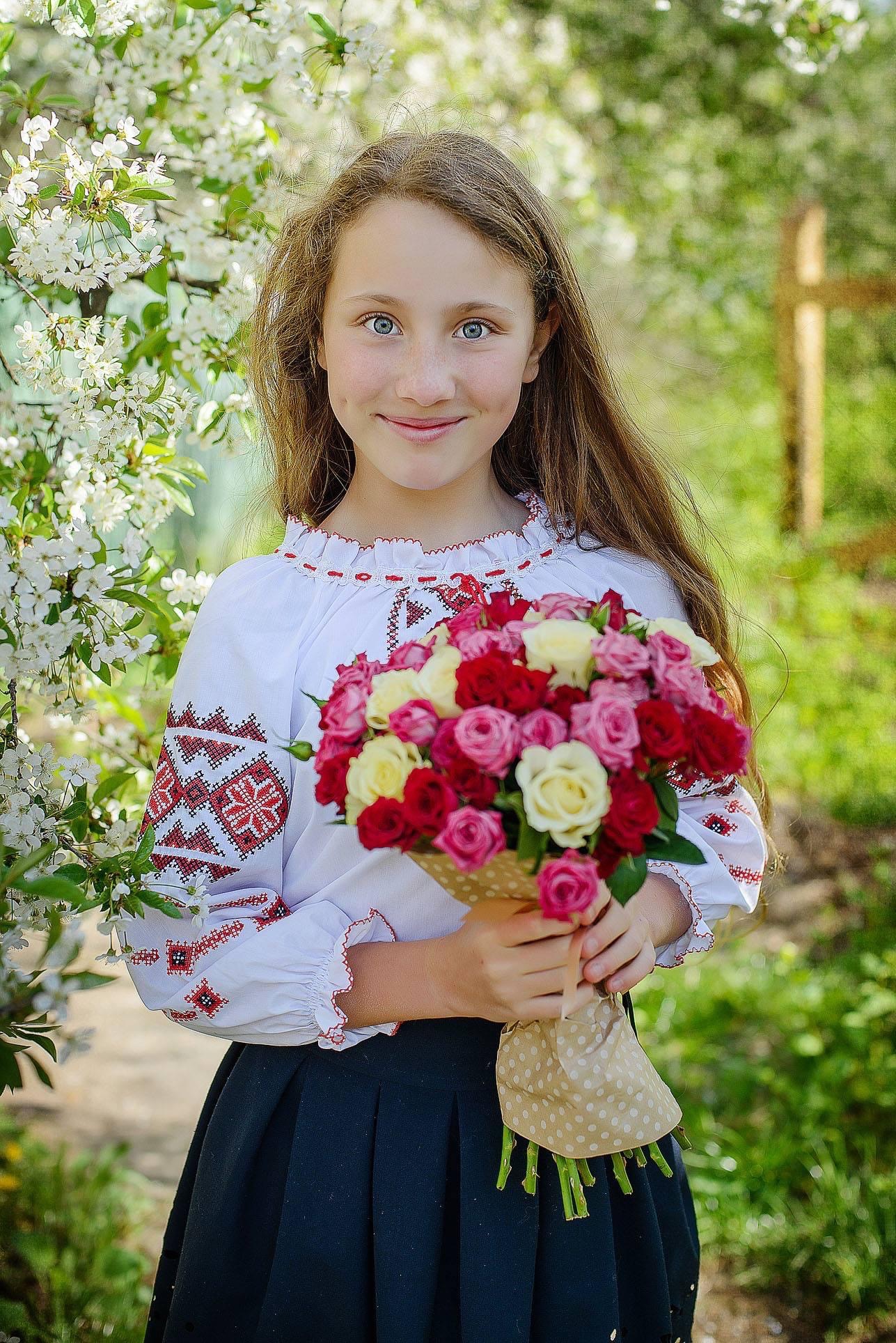 Dziewczynka w haftowanej bluzce trzyma bukiet róż w papierowym opakowaniu na tle kwitnącego drzewa.