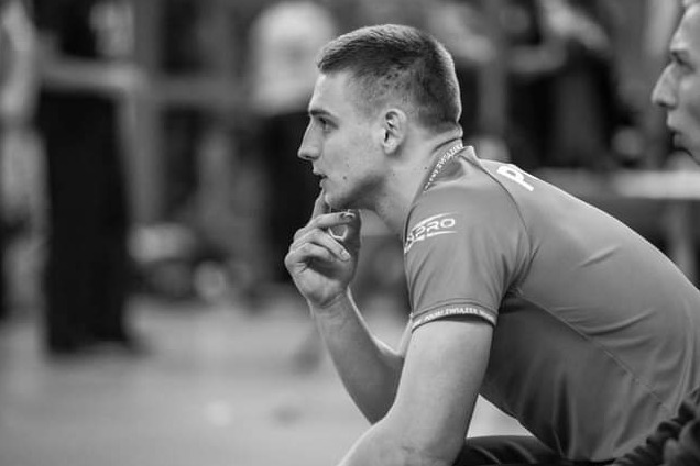 Czarno-białe zdjęcie profilowe zamyślonego mężczyzny w koszulce sportowej z napisem 'PRO' na rękawie, siedzącego na tle rozmytej publiczności.