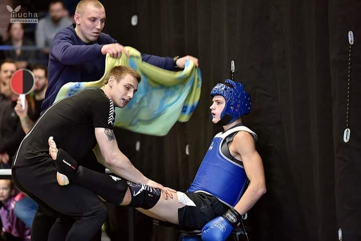 Zawodnik w niebieskim ochraniaczu i rękawicach siedzi na krześle, podczas gdy trener rozciąga mu nogę; w tle drugi trener trzyma ręcznik.