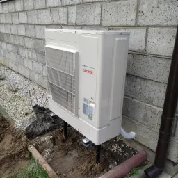 Biała jednostka zewnętrzna z logo Atlantic DC Inverter, zamontowana na metalowych podporach przy szarej ścianie z bloczków betonowych, widoczne elementy instalacji i fragment rury spustowej.