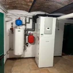 Biała pompa ciepła marki Stiebel Eltron w piwnicy z ceglanym sklepieniem, obok biały zbiornik z niebieskim i czerwonym naczyniem przeponowym, rury w izolacji.