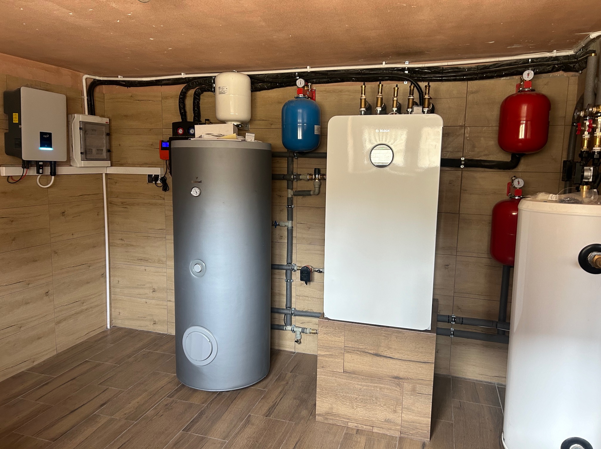 Kotłownia z szarym zasobnikiem ciepłej wody, białym kotłem grzewczym Bosch, czerwonymi i niebieskimi zbiornikami wyrównawczymi, oraz falownikiem fotowoltaicznym FoxESS na ścianie z płytek...