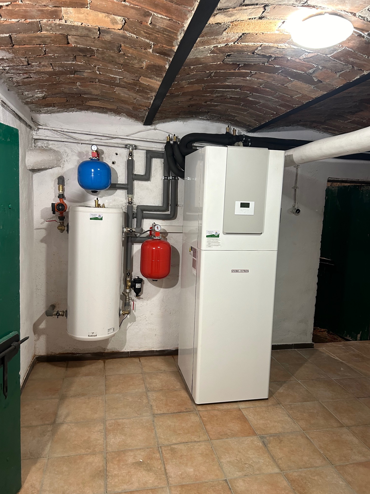 Biała pompa ciepła marki Stiebel Eltron w piwnicy z ceglanym sklepieniem, obok biały zbiornik z niebieskim i czerwonym naczyniem przeponowym, rury w izolacji.
