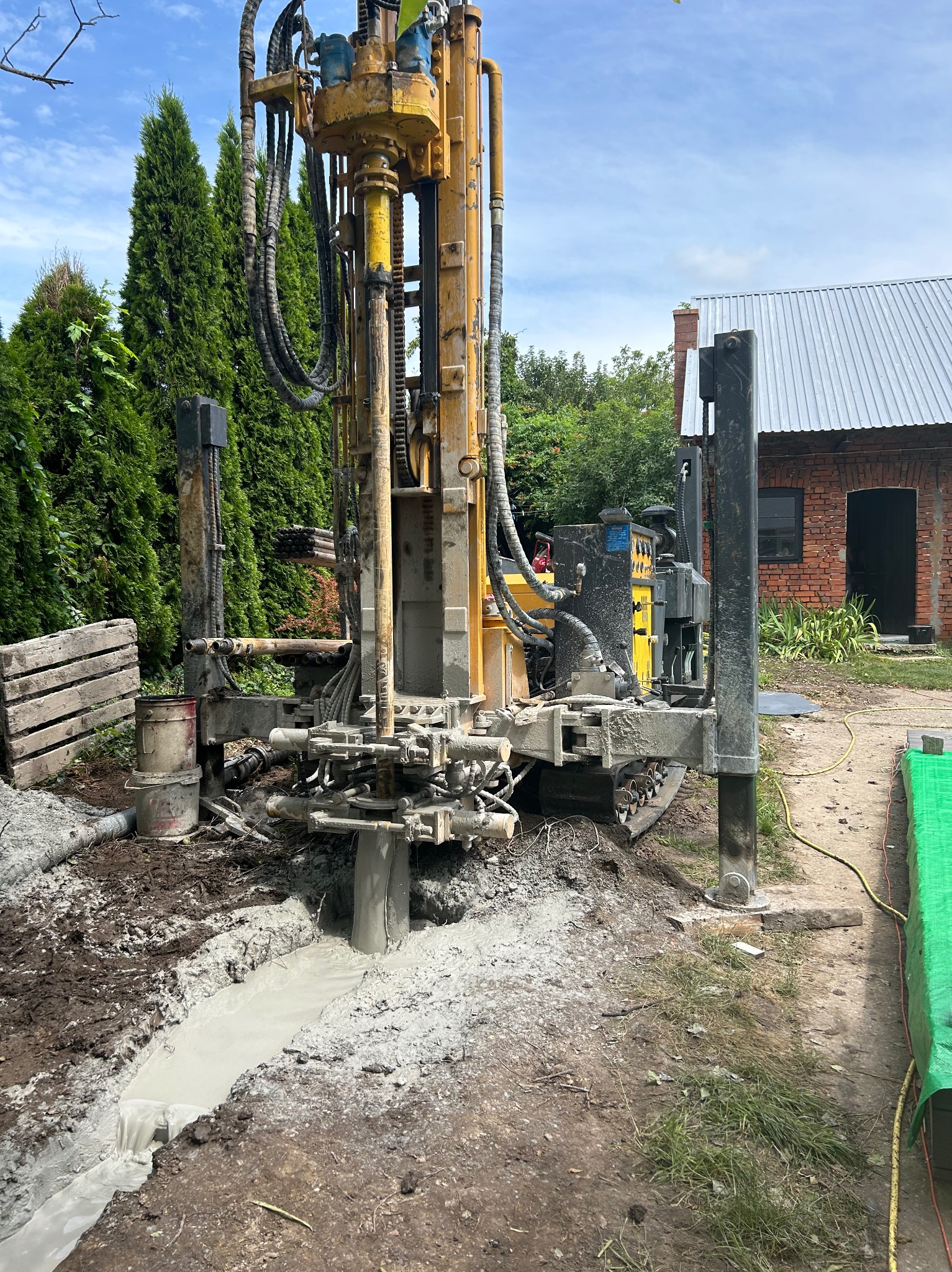 Żółta wiertnica w trakcie wiercenia studni głębinowej, widoczny szlam w wykopie, w tle budynek gospodarczy z blachy falistej i iglaki.