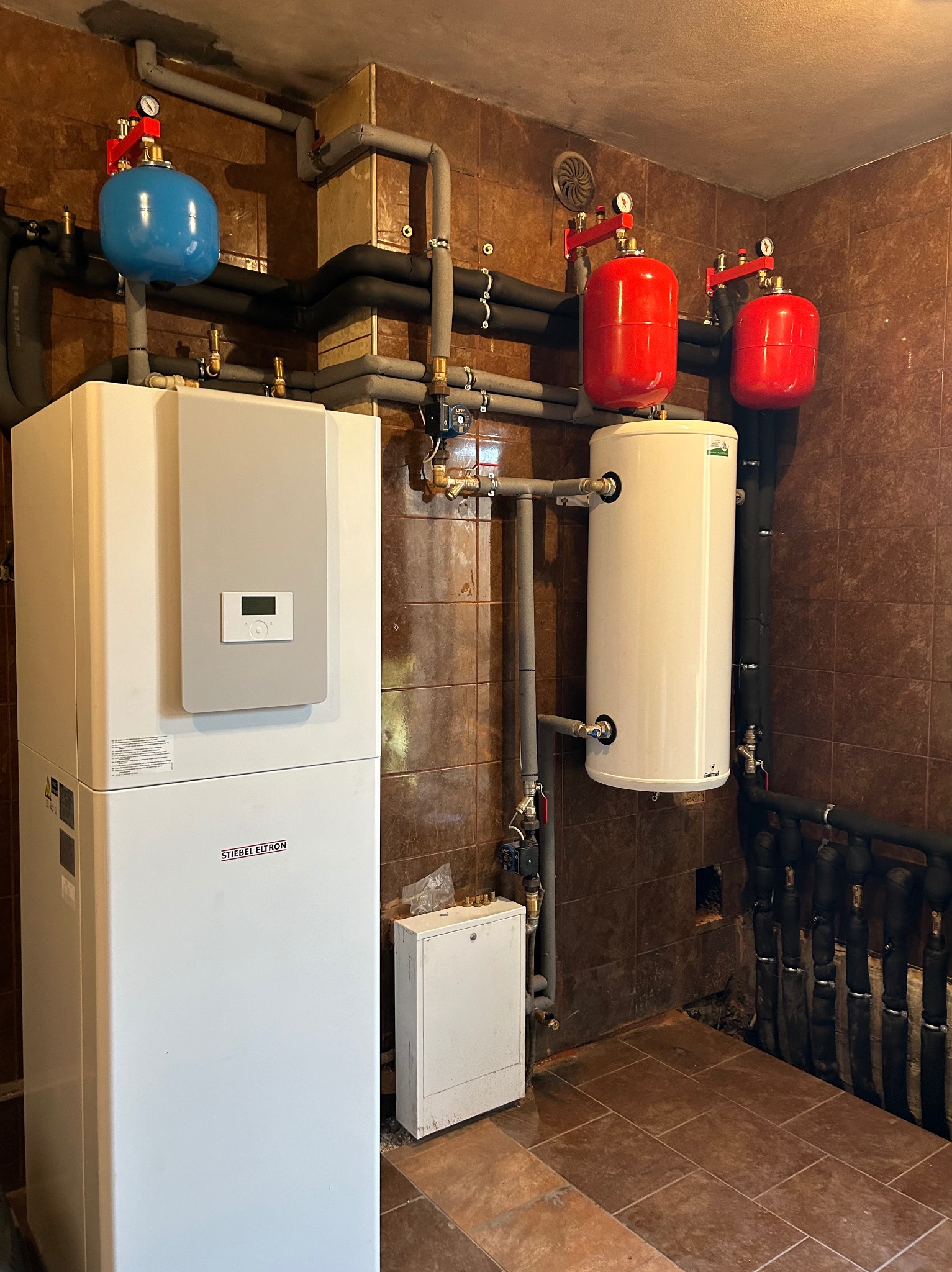 Biała jednostka wewnętrzna z szarą płytą sterującą marki Stiebel Eltron, obok biały zasobnik ciepłej wody z dwoma czerwonymi zbiornikami wyrównawczymi, niebieski zbiornik wyrównawczy, rury...