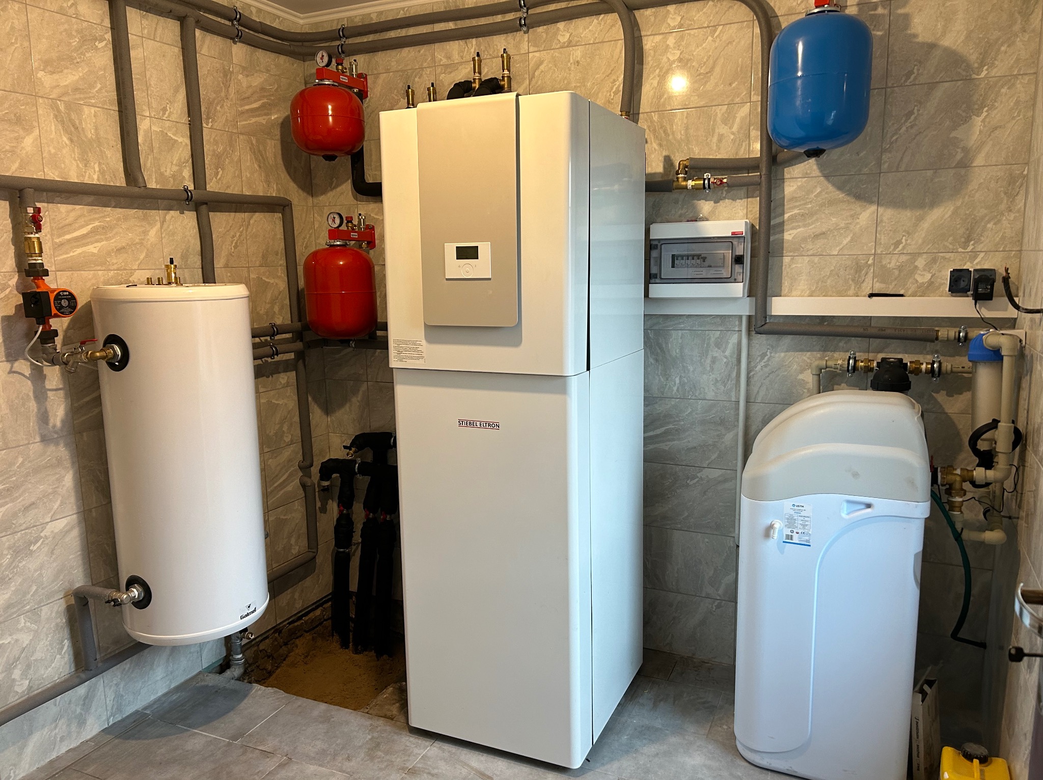 Kompletna instalacja w kotłowni: zbiornik buforowy Galmet, centrala grzewcza Stiebel Eltron, zmiękczacz wody USTH oraz dwa czerwone i jeden niebieski zbiornik wyrównawczy. Widoczne liczne rury...