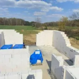 Wznoszone ściany parteru z białych bloczków, zabezpieczone niebieską folią, widok z góry na tle zielonego pola i lasu.