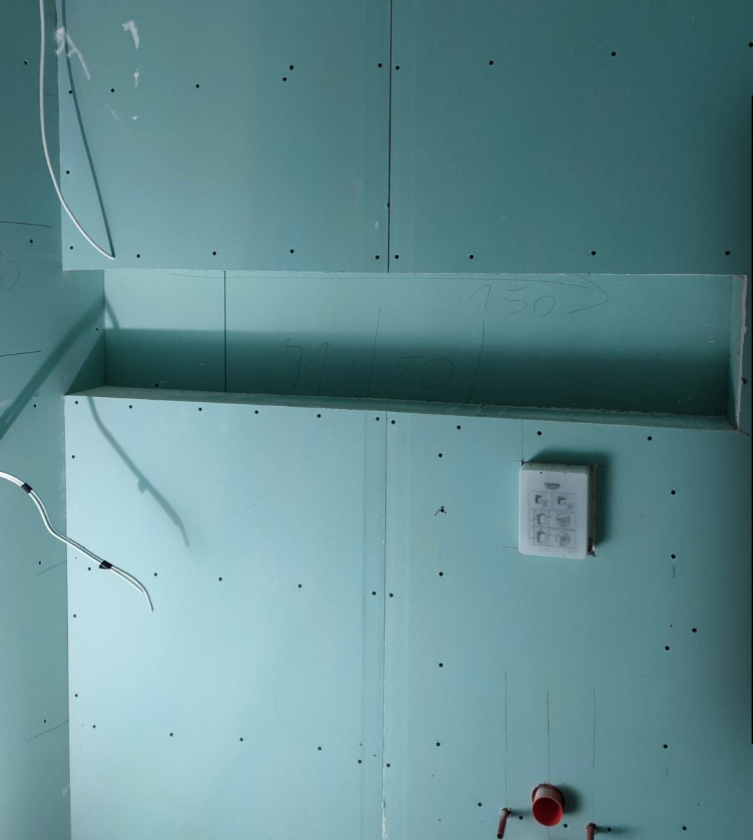 Niebiesko-zielone płyty gipsowo-kartonowe na ścianie z wnęką, widoczne ślady ołówka z wymiarami, instalacja elektryczna i hydrauliczna w trakcie montażu.