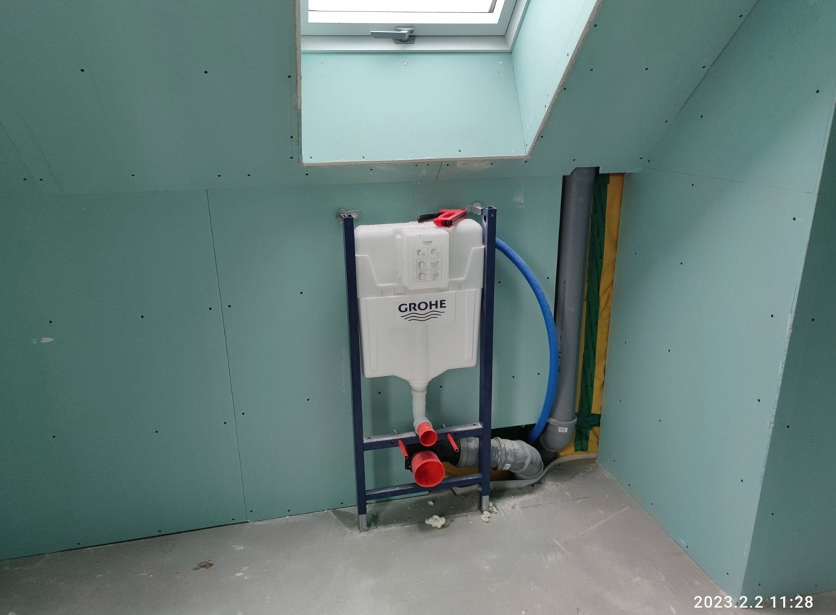 Stelaż podtynkowy Grohe zamontowany przy ścianie z płyty karton-gips w łazience na poddaszu ze skośnym sufitem i oknem dachowym, widoczne elementy instalacji wodno-kanalizacyjnej.