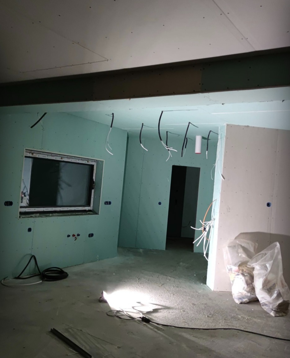 Wnętrze w trakcie prac wykończeniowych: ściany obłożone płytami gipsowo-kartonowymi, widoczne kable elektryczne wystające ze ścian i sufitu, oraz rozproszone światło lampy budowlanej na podłodze.