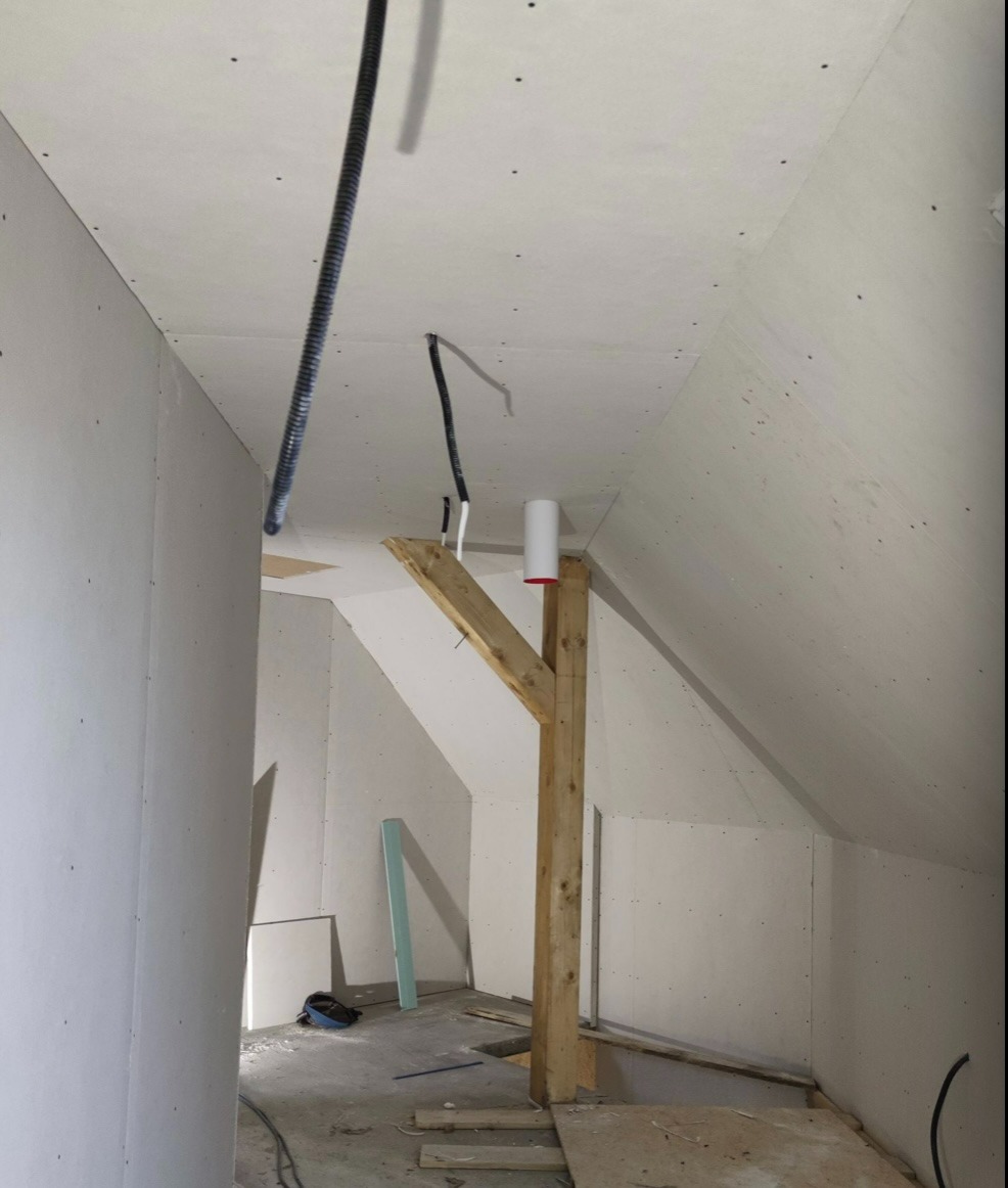 Poddasze w trakcie wykańczania płytami gipsowo-kartonowymi, widoczna drewniana konstrukcja wsporcza, wystające przewody elektryczne oraz fragment instalacji wentylacyjnej.