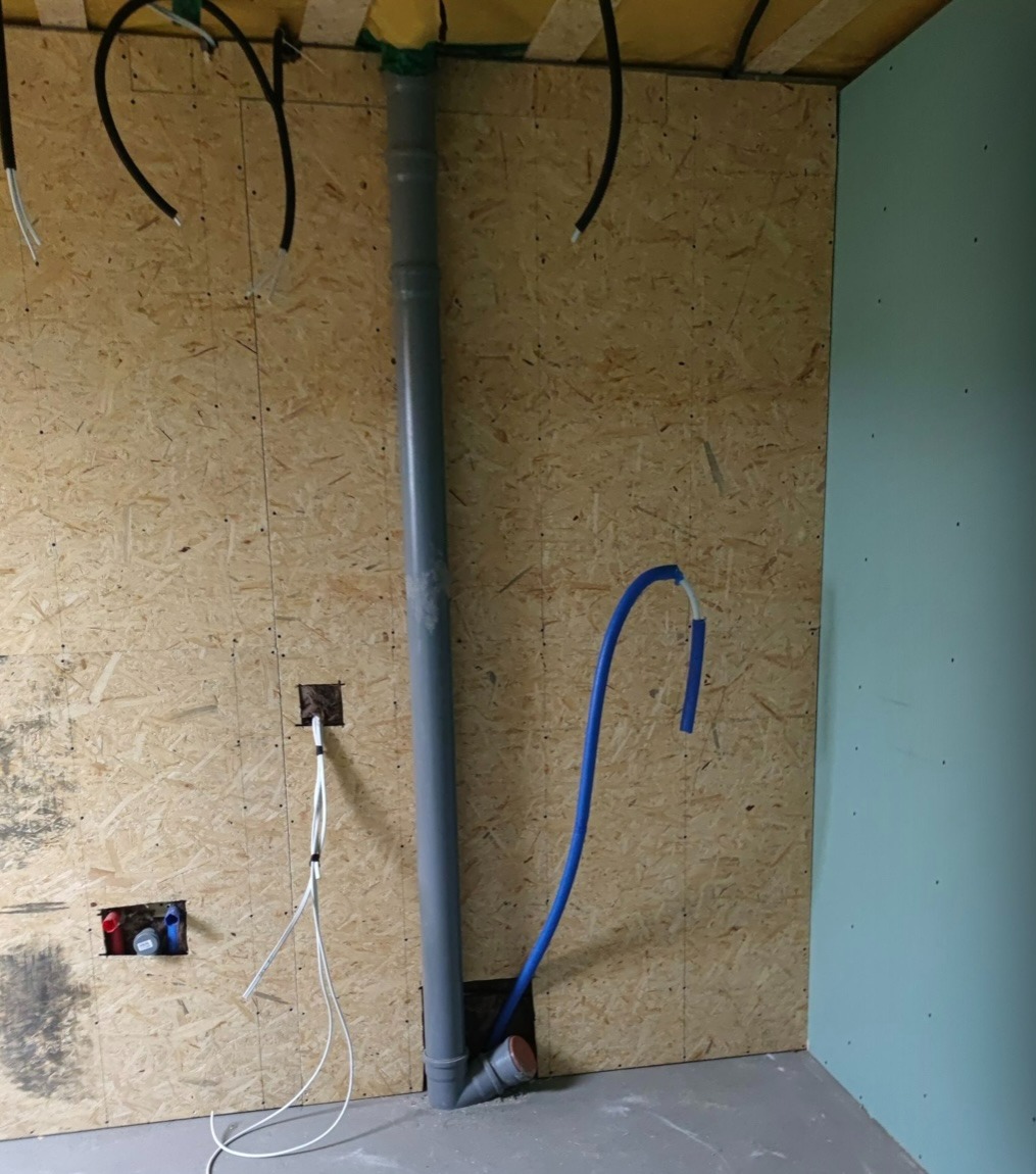Wnętrze w trakcie prac wykończeniowych: ściana z płyty OSB z instalacją wodno-kanalizacyjną i elektryczną, widoczna pionowa rura kanalizacyjna, niebieski przewód instalacji wodnej oraz przewody...