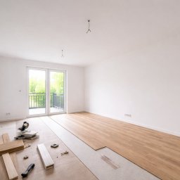Interiora - Jasne wnętrze z częściowo ułożoną podłogą z paneli, narzędzia i deski leżą na podłożu. Widoczne okno balkonowe i białe ściany.