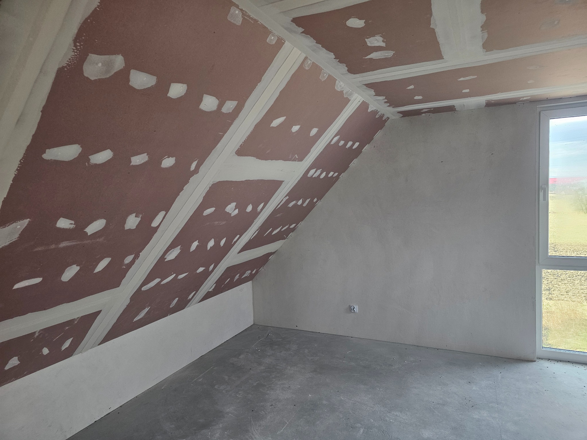 Poddasze w Gorzycach w trakcie prac wykończeniowych: widoczne surowe ściany, płyty na skosach, szpachlowane łączenia i gruntowany beton na podłodze, okno w białej ramie.
