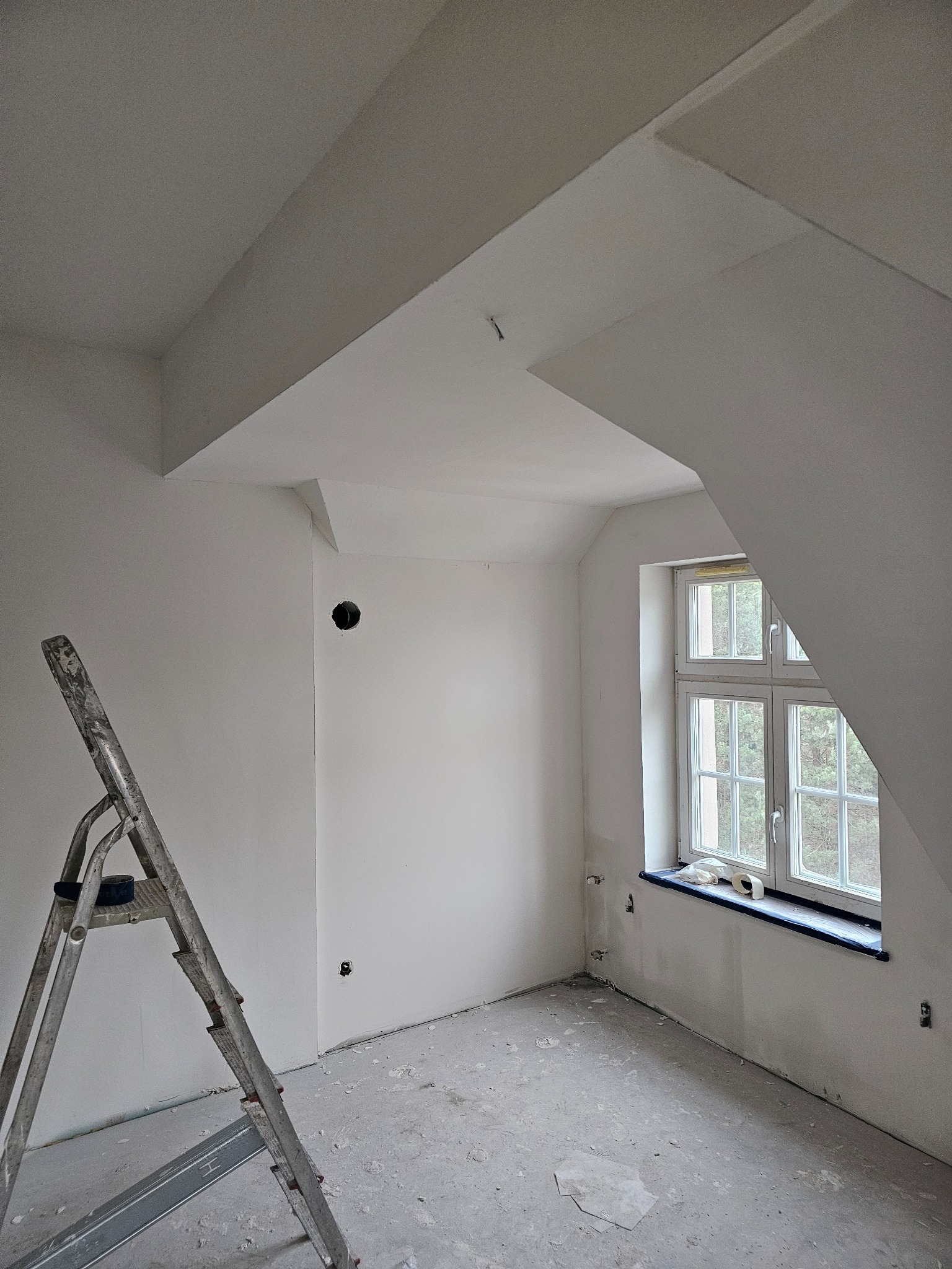 Pokój na poddaszu po świeżym malowaniu na biało, widoczna drabina malarska, okno z parapetem zabezpieczonym folią, gładkie ściany z przygotowanymi otworami na gniazdka.