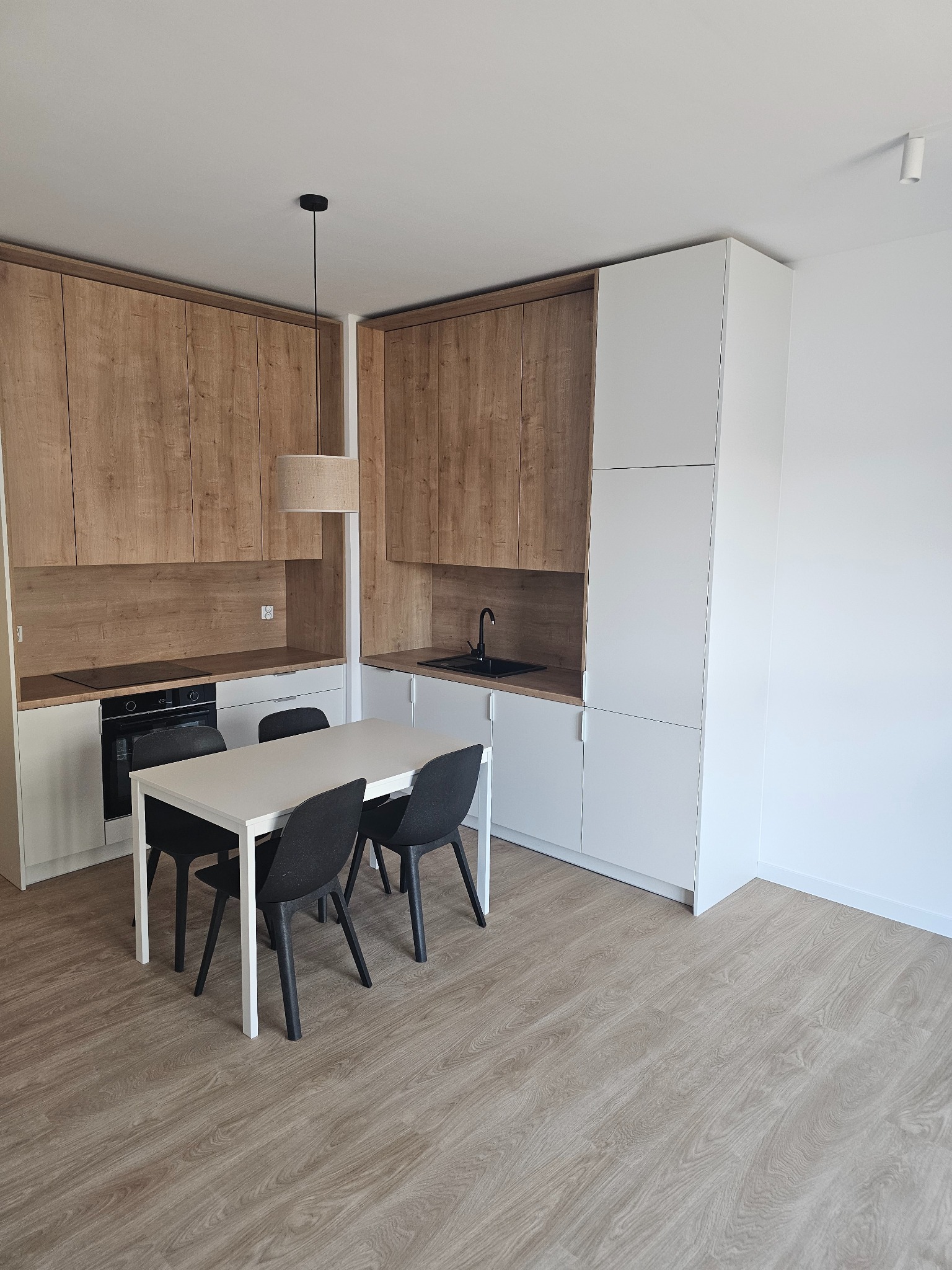 Nowoczesna kuchnia z drewnianymi i białymi szafkami, czarnym zlewem i płytą indukcyjną, biały stół z czarnymi krzesłami, podłoga z paneli imitujących drewno, minimalistyczna lampa wisząca.