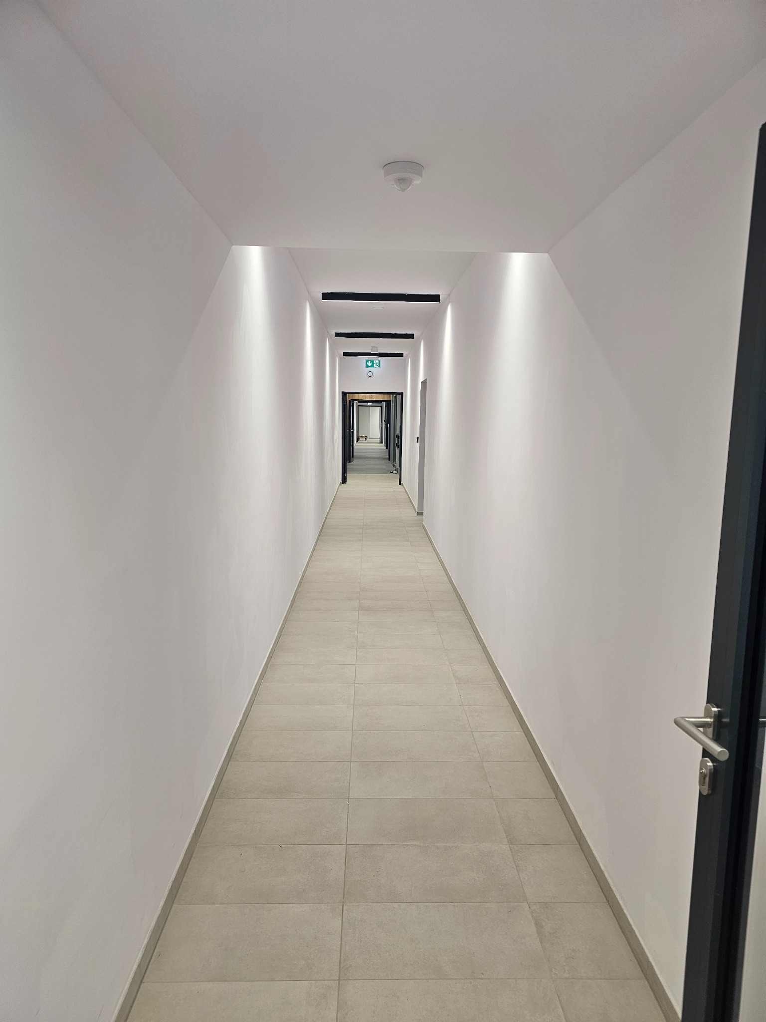 Długi, biały korytarz z szarymi płytkami na podłodze, oświetlony punktowymi lampami w suficie i zakończony drzwiami ewakuacyjnymi z zielonym znakiem.