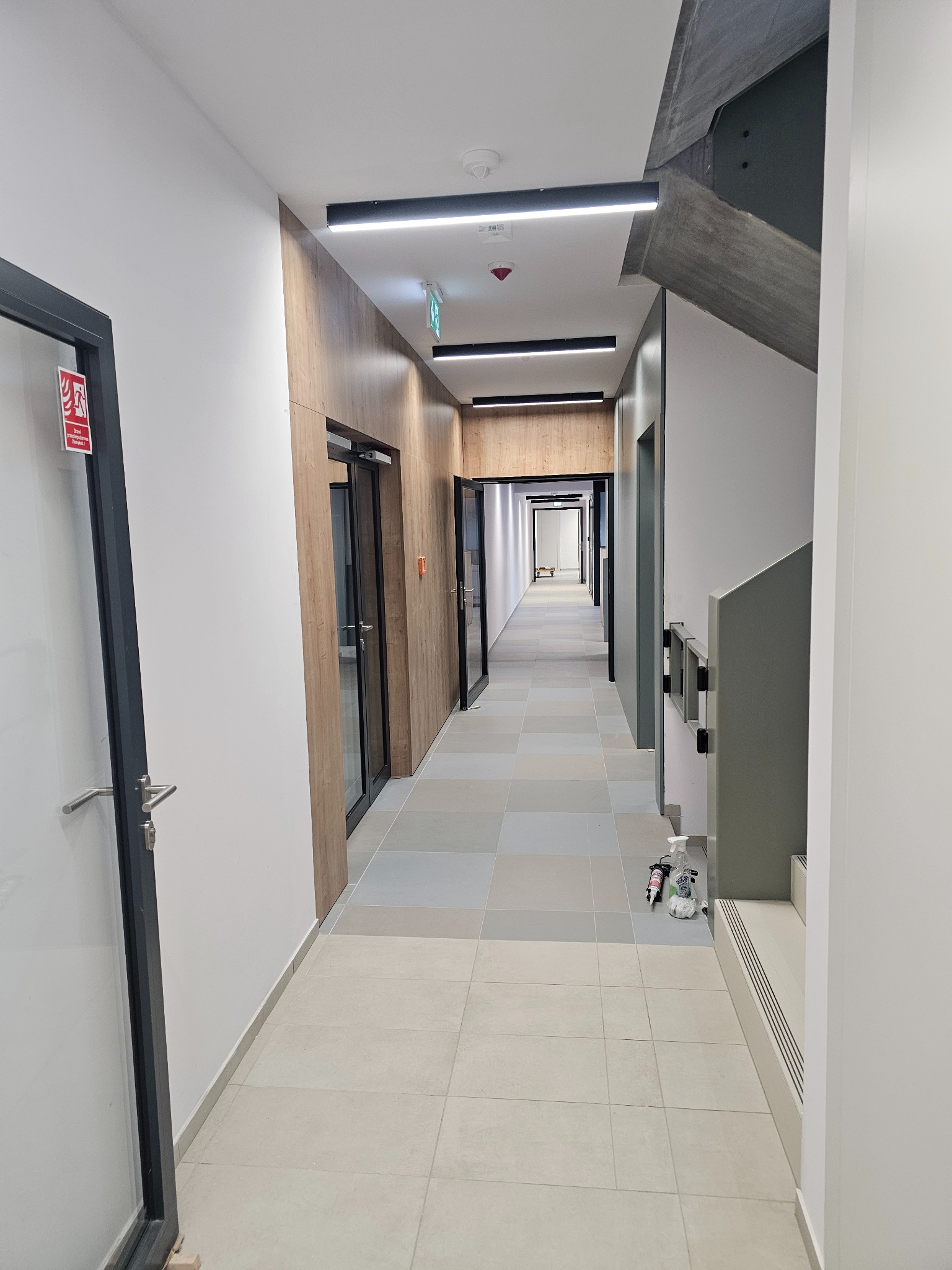 Długi korytarz w budynku biurowym z panelami drewnopodobnymi na ścianach, szarą stolarką drzwiową, płytkami podłogowymi w odcieniach szarości i białymi ścianami, oświetlony liniowymi lampami LED,...