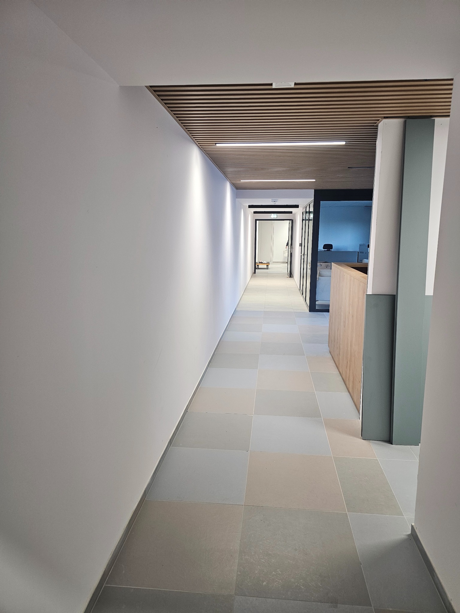 Wnętrze korytarza biurowego z jasnymi ścianami, płytkami podłogowymi w odcieniach szarości i beżu, drewnianą zabudową sufitu oraz widoczną recepcją w tle.