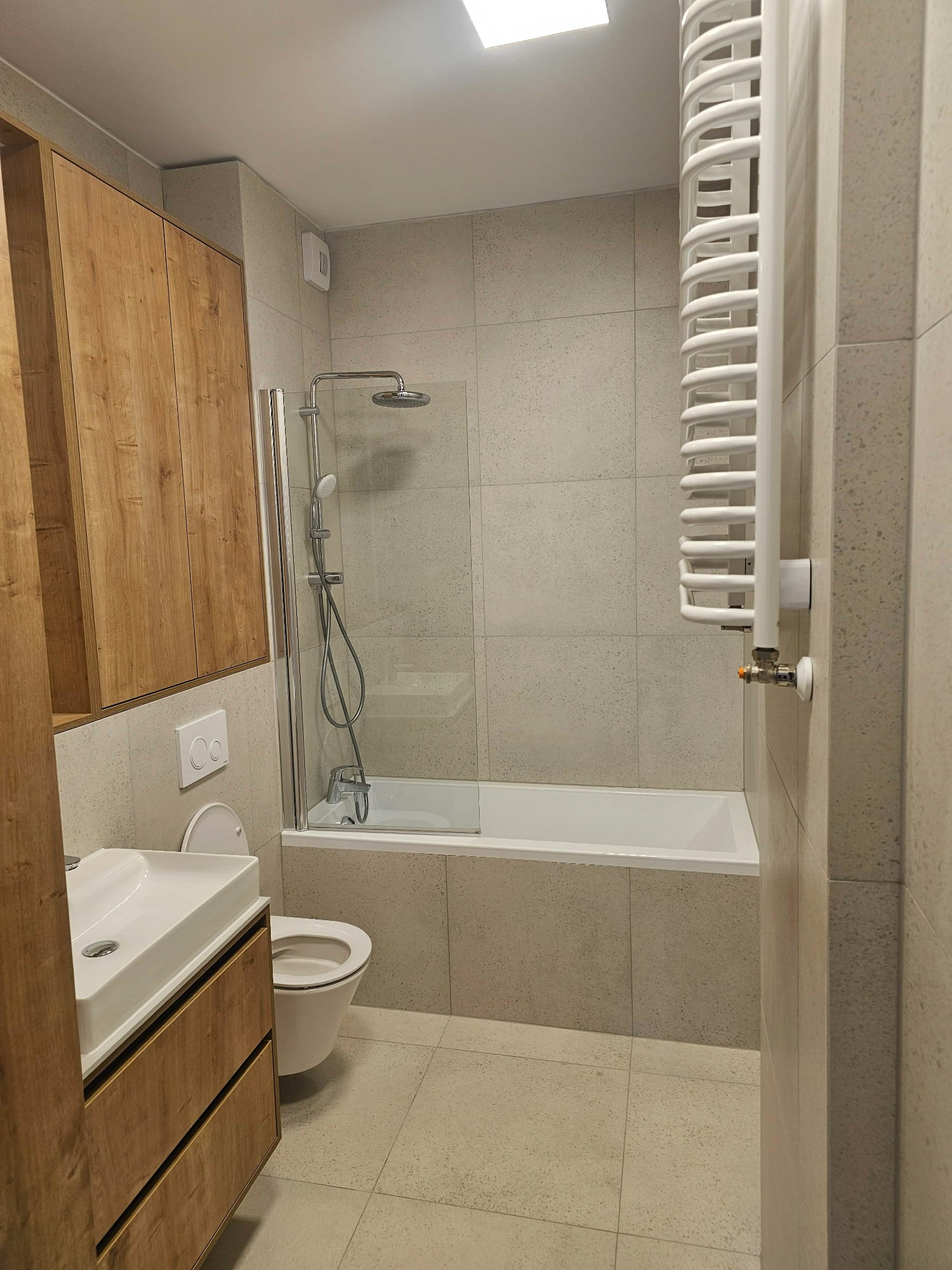 Nowoczesna łazienka w beżowej tonacji: drewniana szafka pod umywalką, biała wanna z parawanem, grzejnik drabinkowy i toaleta podtynkowa. Minimalistyczny design.