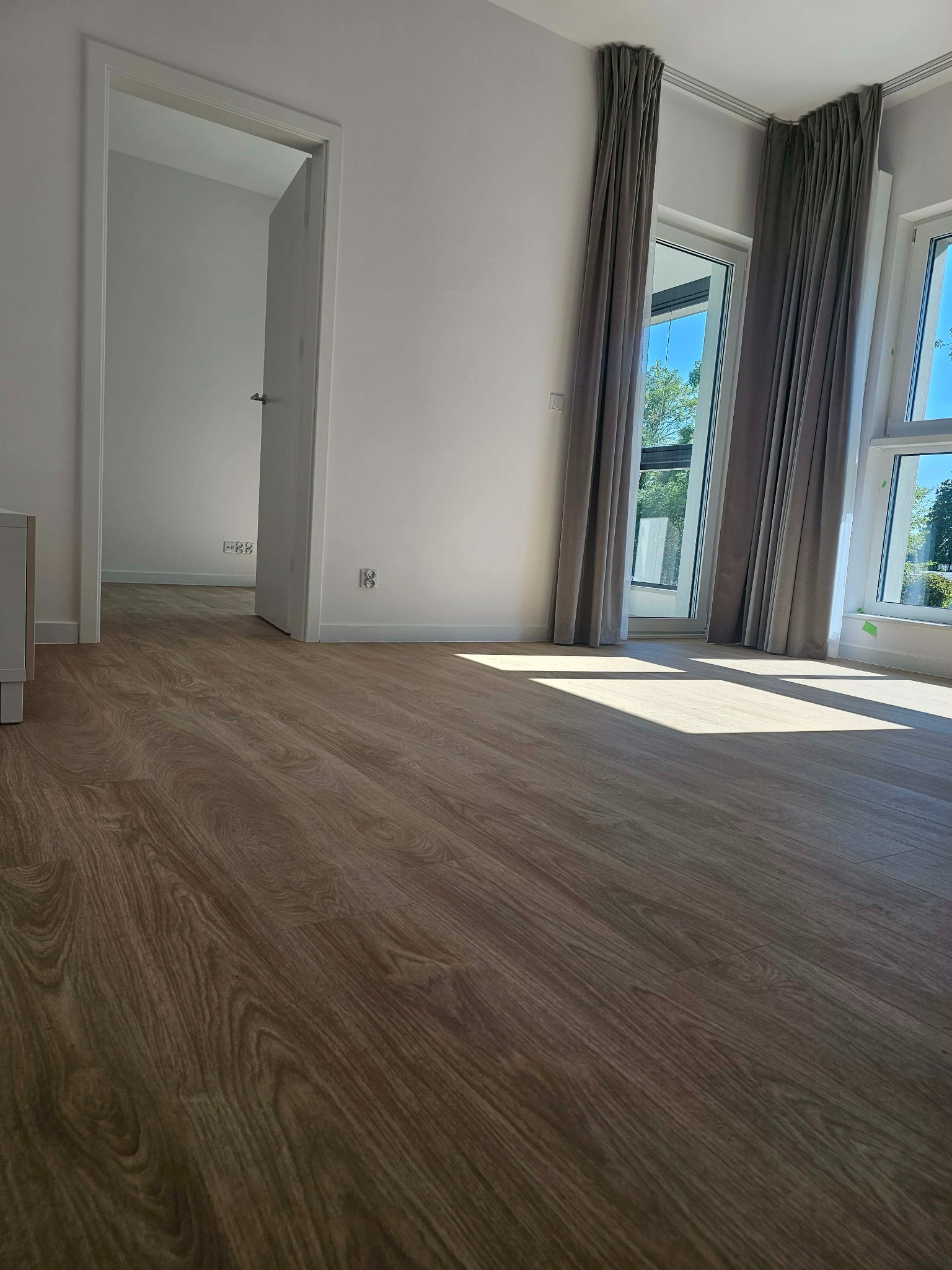 Jasne, przestronne wnętrze z drewnianą podłogą, dużymi oknami z szarymi zasłonami i otwartymi drzwiami do kolejnego pomieszczenia. Wpadające światło słoneczne.