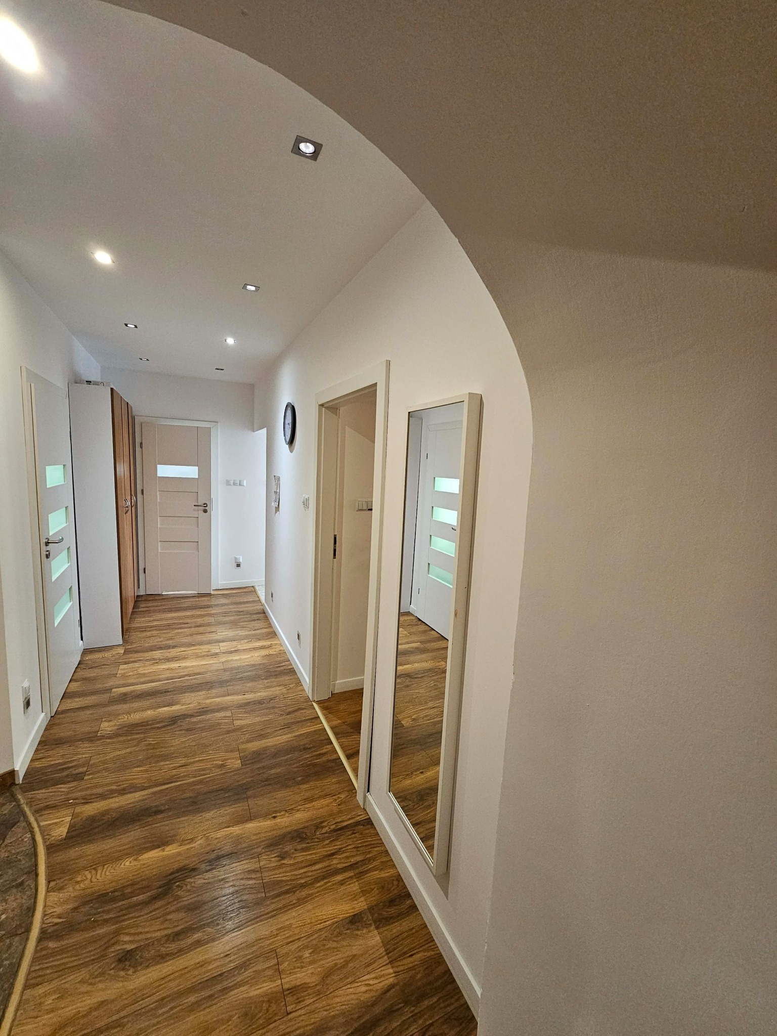 Korytarz po remoncie: drewnopodobna podłoga, białe ściany z łukiem, lustro w ramie, drzwi z przeszkleniami i punktowe oświetlenie sufitowe. Widok w głąb.