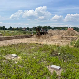 Przygotowanie terenu pod fundamenty: wykop, szalunki z desek, pracujący traktor z łyżką, hałdy ziemi, widok na okoliczne domy i błękitne niebo z chmurami.
