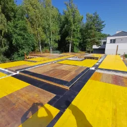 Żółte szalunki stropowe z widocznym zbrojeniem na tle drzew i nowoczesnego domu w trakcie budowy.