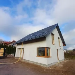 Surowy, otynkowany dom jednorodzinny z ciemną dachówką i drewnianymi oknami, widok z zewnątrz, z widoczną rurą doprowadzającą materiał i samochodem dostawczym w tle.