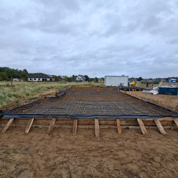 LW CONSTRUCTION S.C. CEZARY WAŚNIEWSKI, KRZYSZTOF LEŚKIEWICZ - Zbrojenie fundamentu pod budowę domu jednorodzinnego, widok na siatkę zbrojeniową w drewnianym szalunku, przygotowaną do zalania betonem na tle pochmurnego nieba.