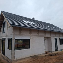 LW CONSTRUCTION S.C. CEZARY WAŚNIEWSKI, KRZYSZTOF LEŚKIEWICZ - Niedokończony dom z białych bloczków, czarną dachówką i oknami z roletami zewnętrznymi. Widoczne elementy konstrukcyjne i brak elewacji. Szary dzień.