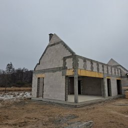 LW CONSTRUCTION S.C. CEZARY WAŚNIEWSKI, KRZYSZTOF LEŚKIEWICZ - Surowy stan domu z białych bloczków, widoczne otwory okienne i drzwiowe, betonowe nadproża, w tle drzewa i częściowo zaśnieżone pole. Budowa w toku.
