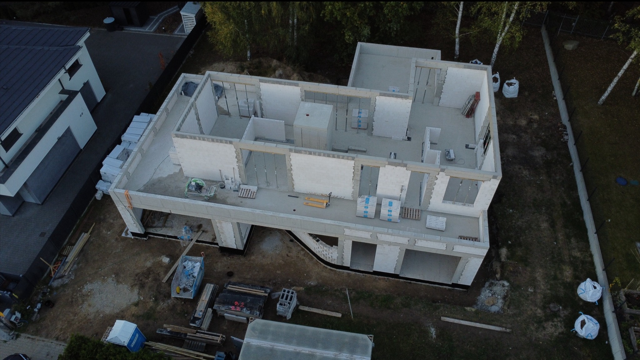 Zdjęcie z drona przedstawia budynek w stanie surowym otwartym z białymi ścianami z betonu komórkowego, widoczne materiały budowlane na stropie oraz palety z bloczkami, teren wokół budynku częściowo...