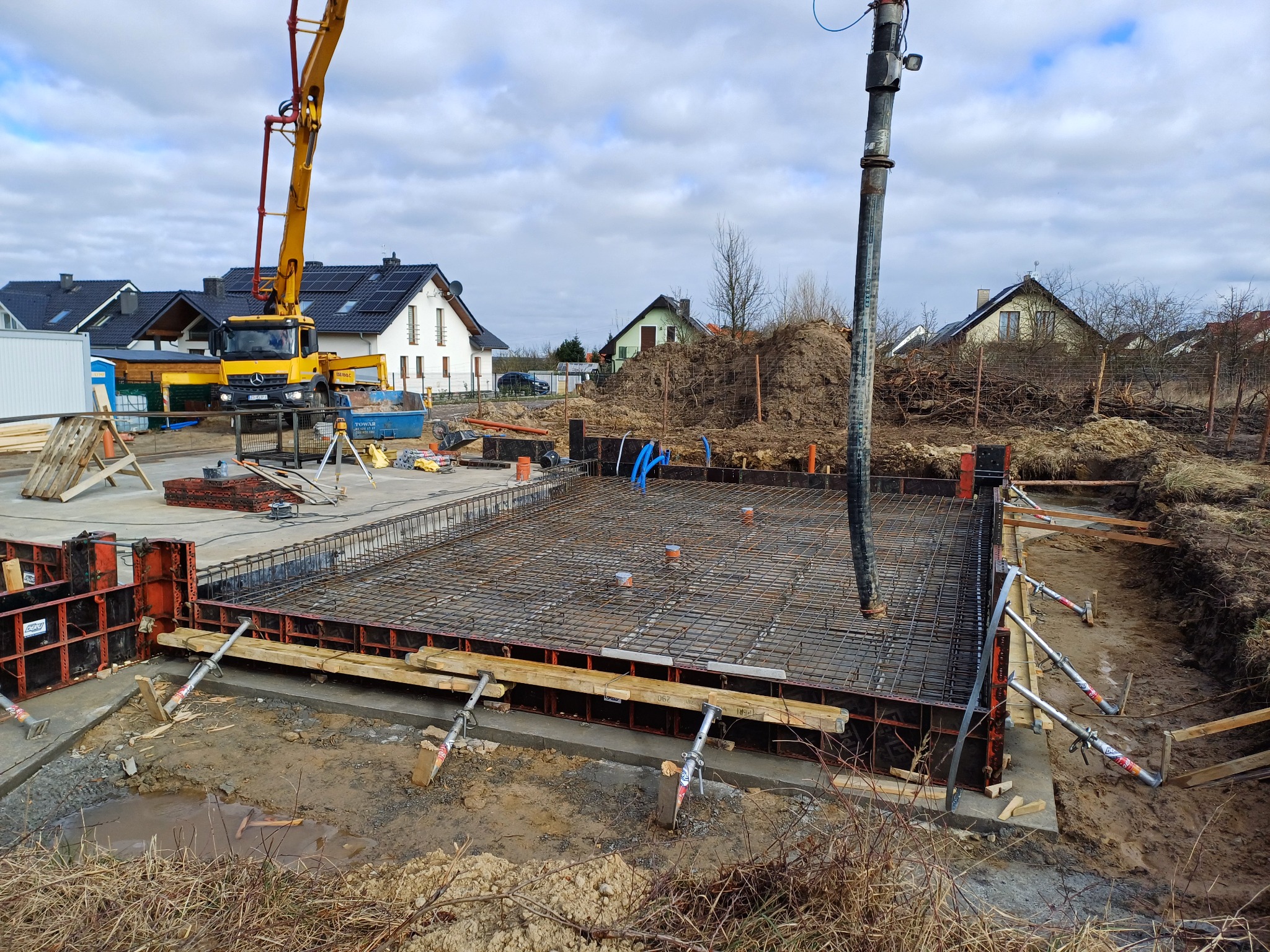 Wylewanie betonu do szalunku zbrojonego na budowie fundamentów domu jednorodzinnego; w tle widoczny samochód z pompą do betonu i zabudowa osiedla.