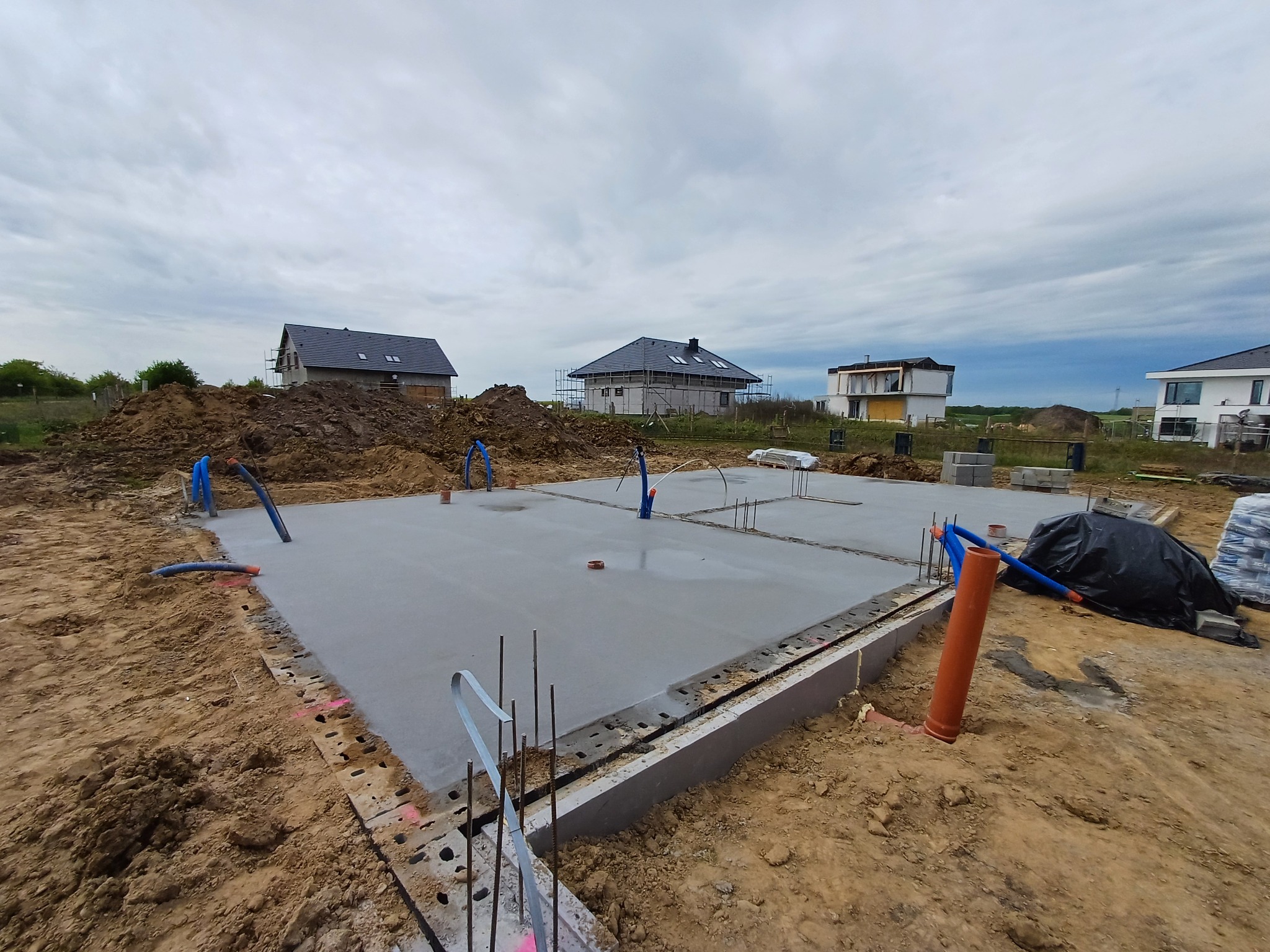 Wylany fundament płyty fundamentowej z wystającymi rurami instalacyjnymi, widoczne zbrojenia, w tle budynki w trakcie budowy.