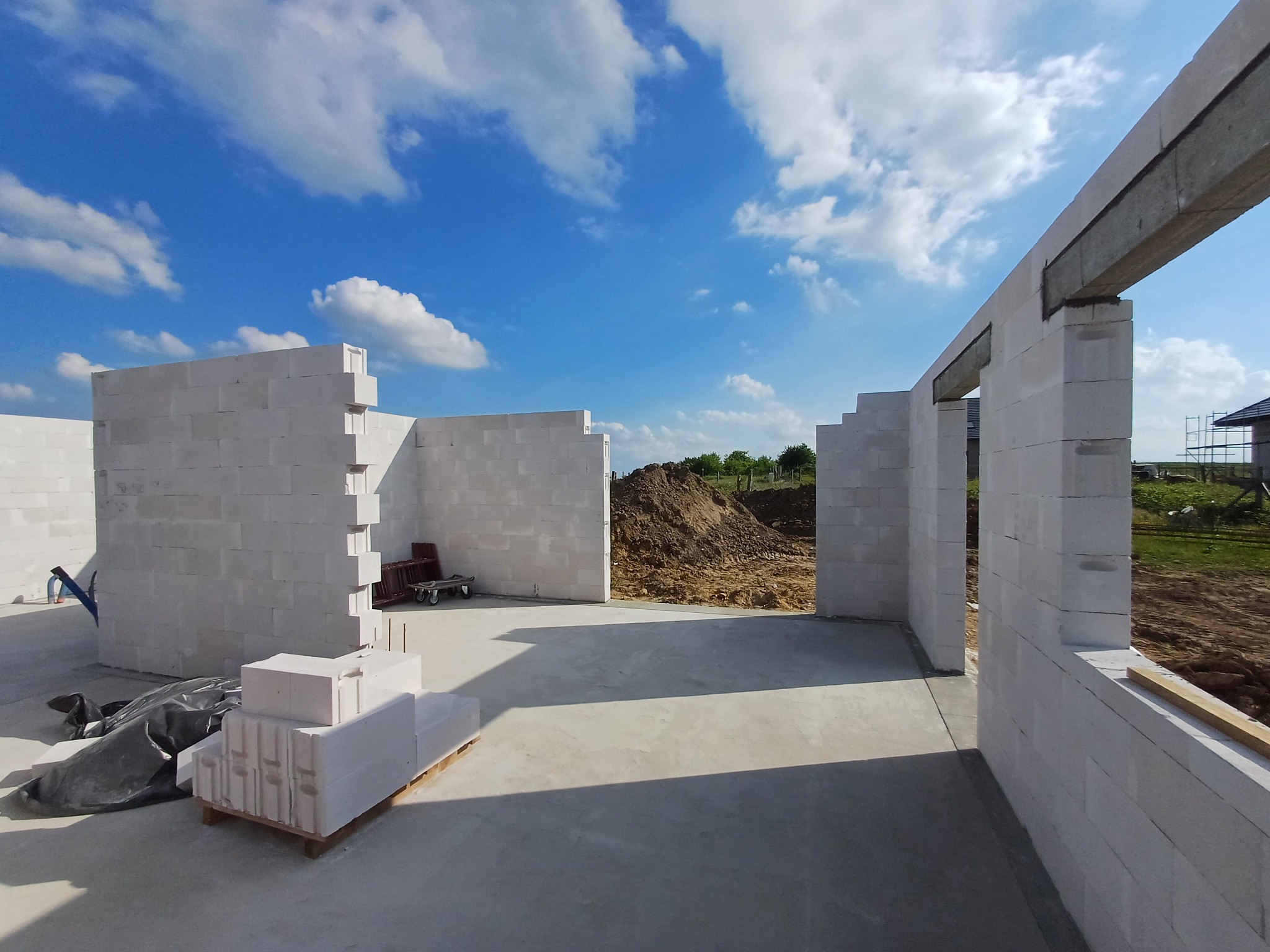 Wnętrze w trakcie budowy domu z białych bloczków, widoczne ściany wzniesione do pewnej wysokości, paleta bloczków i fragment nieba z chmurami w tle.