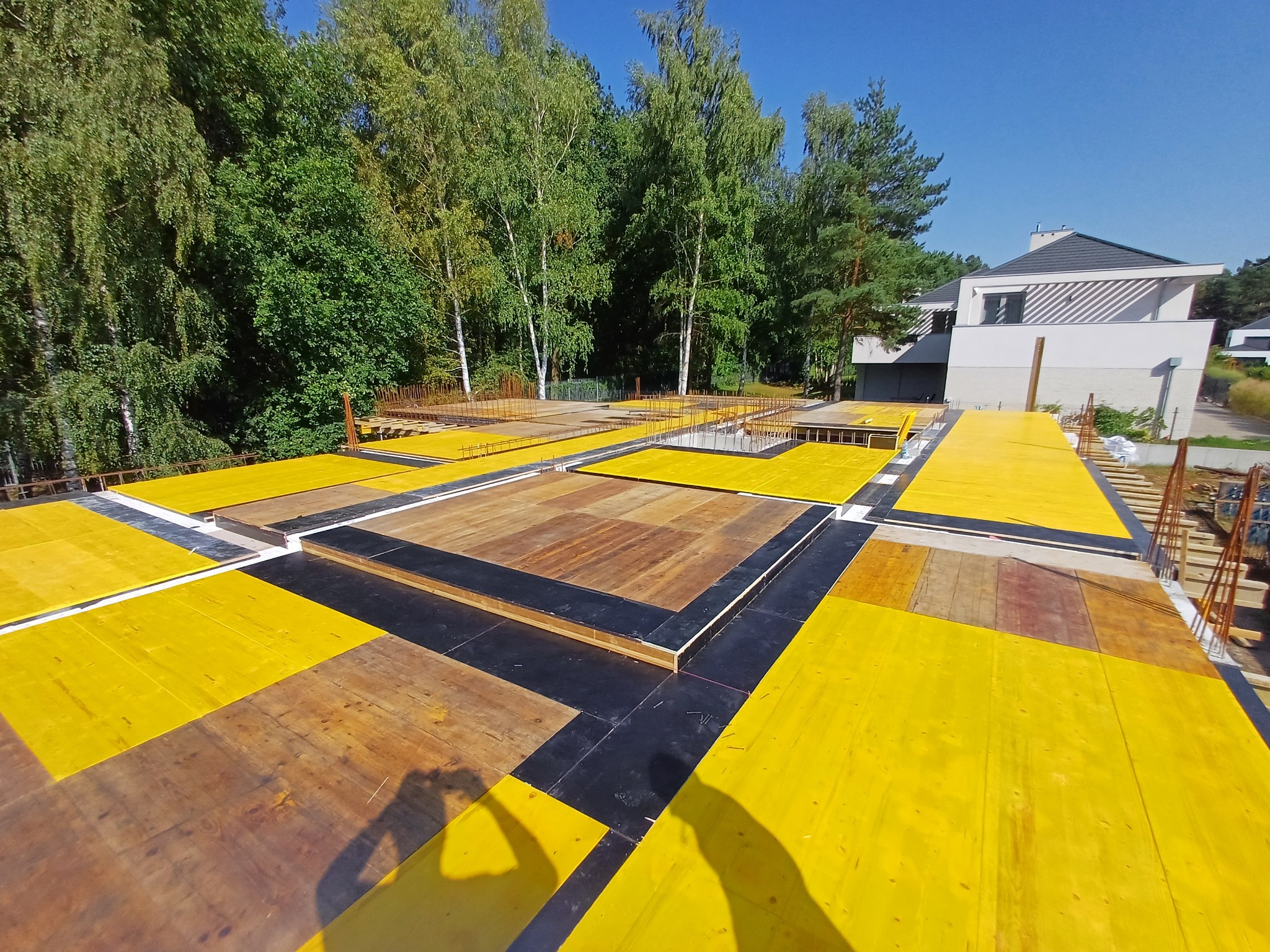 Żółte szalunki stropowe z widocznym zbrojeniem na tle drzew i nowoczesnego domu w trakcie budowy.