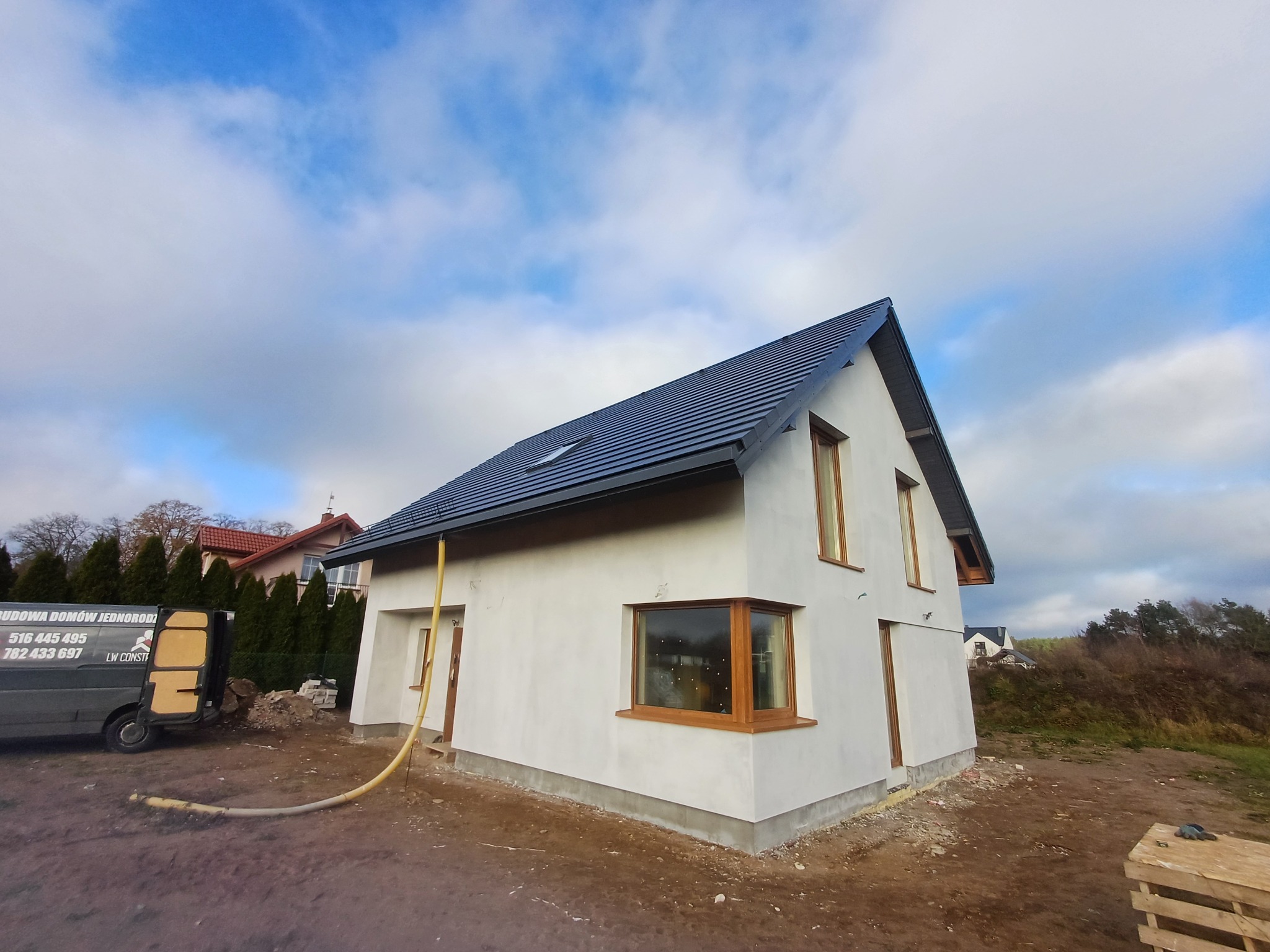 Surowy, otynkowany dom jednorodzinny z ciemną dachówką i drewnianymi oknami, widok z zewnątrz, z widoczną rurą doprowadzającą materiał i samochodem dostawczym w tle.