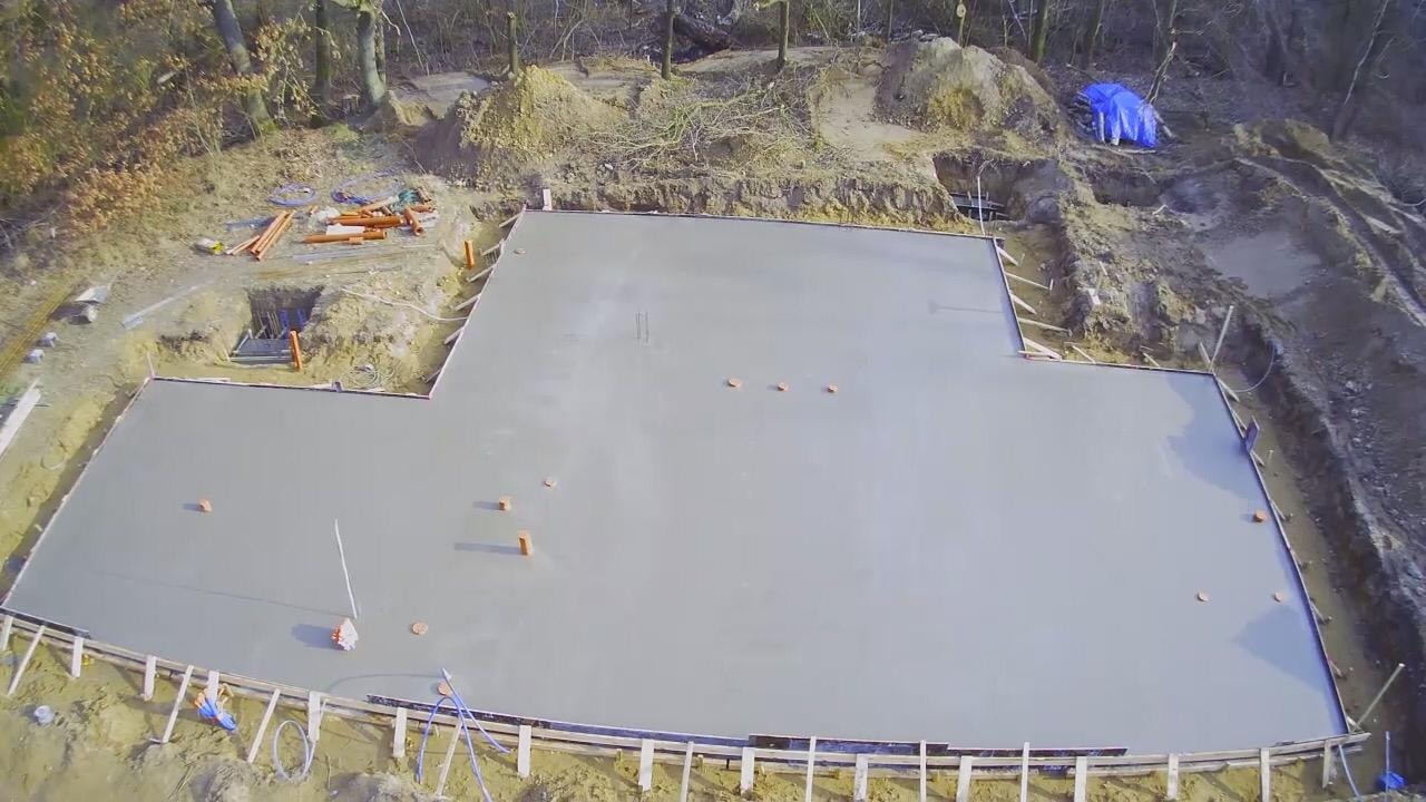 Świeżo wylany, gładki betonowy fundament pod budowę domu jednorodzinnego, widok z góry, z widocznymi elementami szalunku i wystającymi rurami wentylacyjnymi w tle ziemia i fragment lasu.
