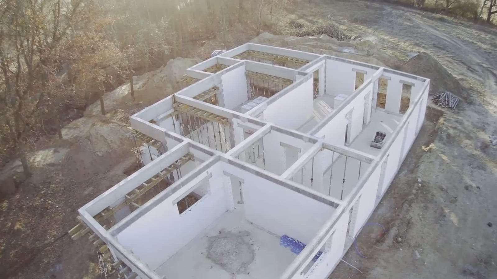 Widok z lotu ptaka na surowy stan domu murowanego z białych bloczków, widoczne drewniane stemple podtrzymujące strop, teren budowy z hałdami ziemi i drzewami w tle.