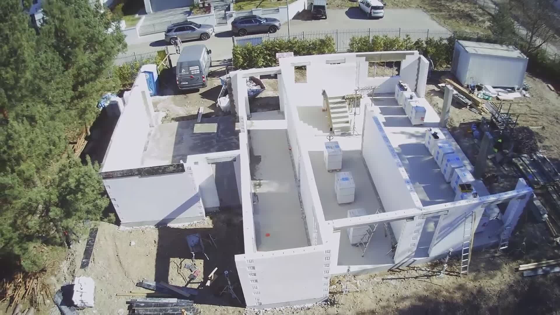 Widok z lotu ptaka na budowę domu z białych bloczków, widoczne fundamenty i wznoszone ściany, na zewnątrz zaparkowane samochody i teren budowy.