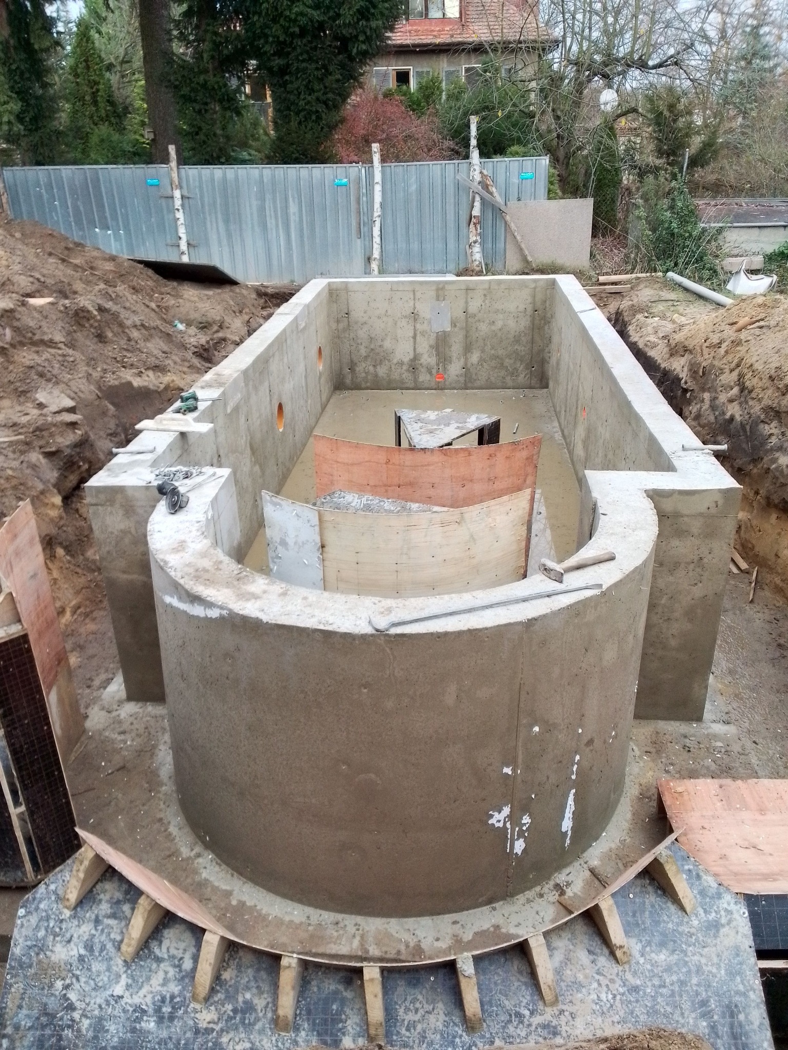 Betonowy fundament z półokrągłym elementem w wykopie, widoczne deski szalunkowe i narzędzia budowlane, ziemia wokół wykopu, w tle ogrodzenie i budynek.