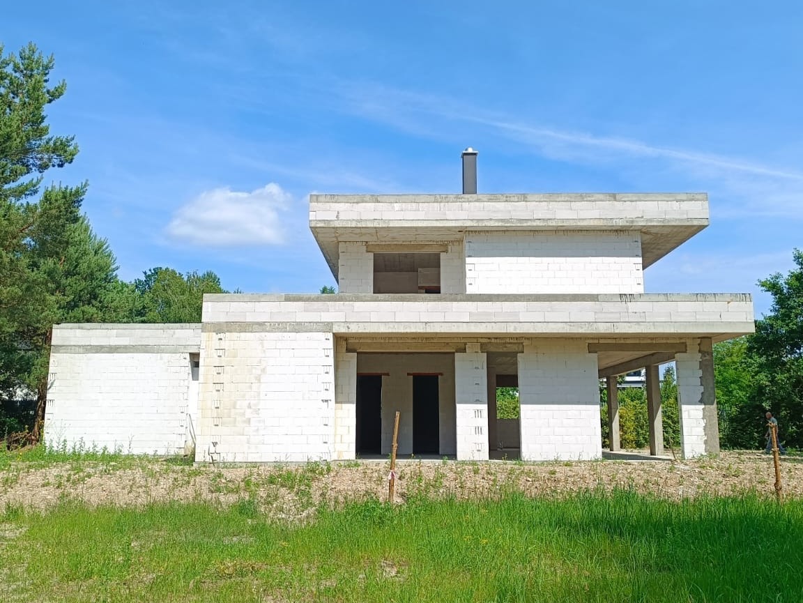 Surowy, dwukondygnacyjny budynek z białych bloczków, z widoczną konstrukcją stropu i otworami okiennymi, w otoczeniu zieleni i błękitnego nieba.